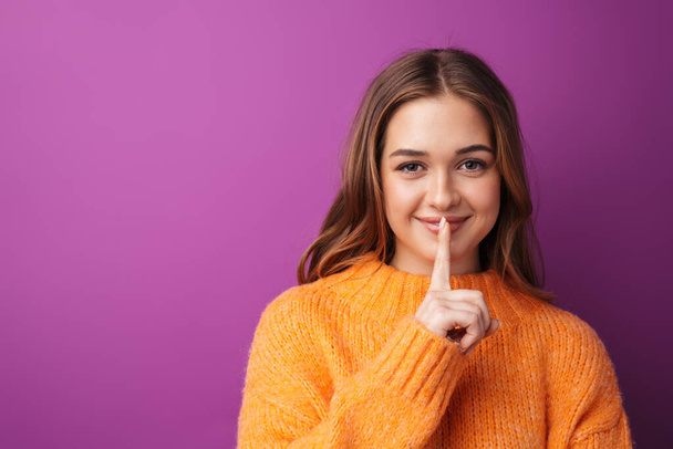 Portrait of a lovely cheerful young girl wearing sweater standing isolated over violet background, showing silence gesture - 写真・画像