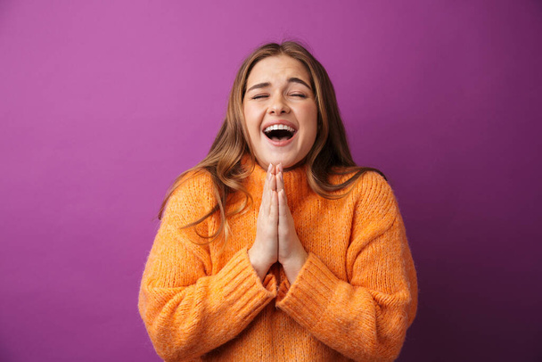 Image of happy young woman wearing sweater smiling and praying with palms together isolated over purple background - Φωτογραφία, εικόνα