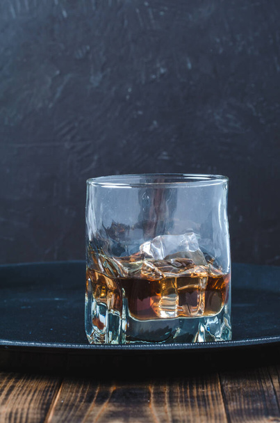 Glass of whiskey with ice cubes on a black tray and wooden table/Glass of whiskey with ice cubes on a black tray and wooden table. Selective focus - Фото, зображення
