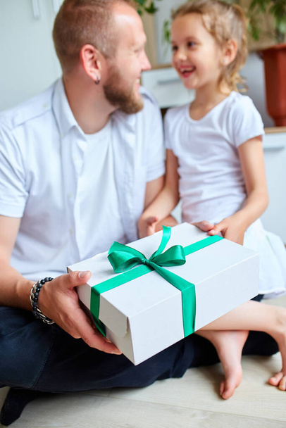 Το κοριτσάκι δίνει στον όμορφο πατέρα της ένα κουτί δώρου την ημέρα του πατέρα, χαμογελαστή κόρη συγχαίροντας τον μπαμπά και δίνοντας δώρο στα γενέθλια στο σπίτι. Σ 'αγαπώ, μπαμπά. Χαρούμενη Ημέρα του Πατέρα. - Φωτογραφία, εικόνα