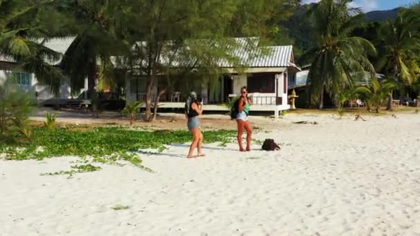 Две юные подружки стоят на песчаном побережье с багажом рядом с ними. Одна девушка фотографирует свою подругу по мобильному телефону. Красивые женщины отдыхают на тропическом курорте - Кадры, видео