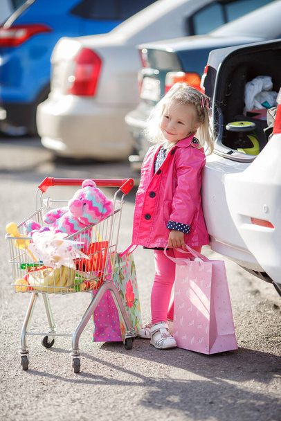 Adorable baby kid with trolley choosing fresh vegetables in local store. Girl with a big grocery cart in a parking lot near a store. A little girl with a big trolley buys fresh food and vegetables in a big supermarket.Little housewife with groceries - 写真・画像
