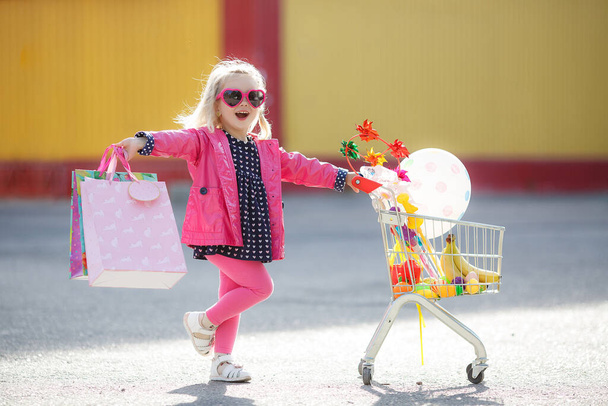 Adorable baby kid with trolley choosing fresh vegetables in local store. Girl with a big grocery cart in a parking lot near a store. A little girl with a big trolley buys fresh food and vegetables in a big supermarket.Little housewife with groceries - Foto, imagen