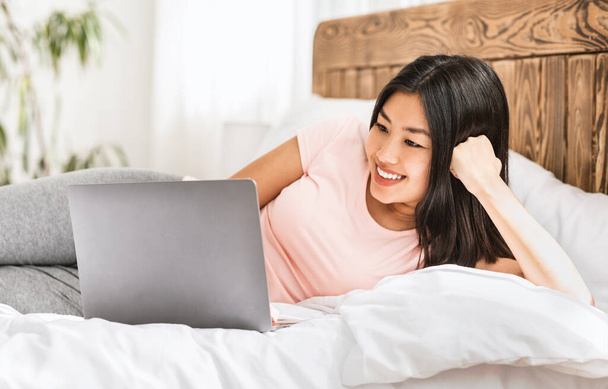 Щаслива азіатська дівчина використовує ноутбук лежить в ліжку вдома
 - Фото, зображення