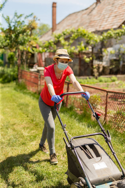 Bahçesinde çim biçme makinesiyle çim biçen bir kadın güneşli bir günde Coronavirus salgını yüzünden cerrahi maske takıyor.. - Fotoğraf, Görsel
