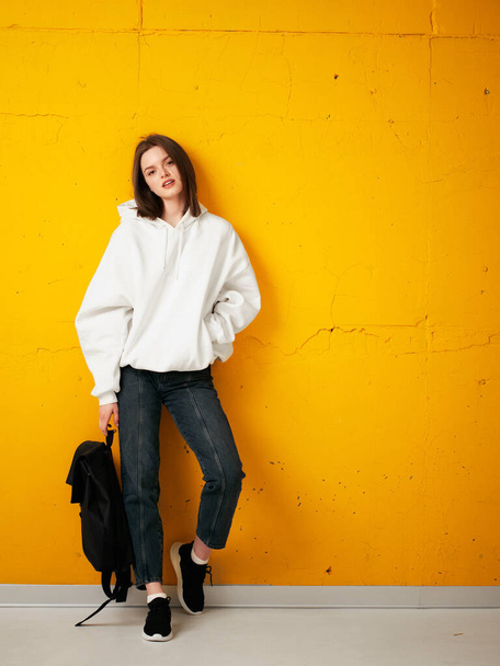 Portrait of a young pretty girl student in a white hoodie, casual clothes, with a bag, against a concrete orange wall. - Photo, image