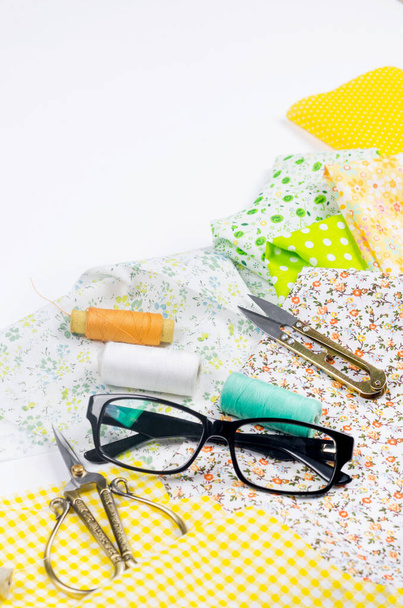 Set of colorful yellow and green fabrics, scissors, buttons, spools of thread and glasses on white. Concept for tailoring, patchwork and handwork. Needlework and Sewing. - Foto, imagen