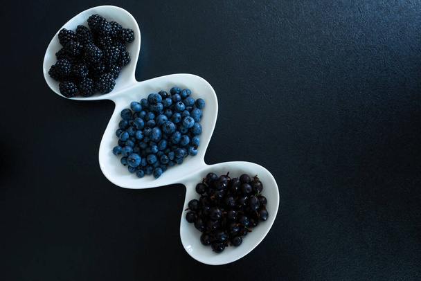 Set of different types of black berries in a white plate on a black table. Stylish seasonal vitamins. - Foto, Imagem
