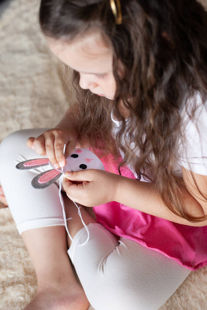 child girl sews a toy made of felt sitting on the bed on a beige blanket; needlework - Fotó, kép