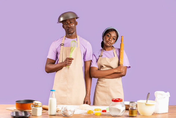 Erschöpftes schwarzes Paar mit Küchenutensilien, die es schwer haben, auf farbigem Hintergrund zu backen - Foto, Bild