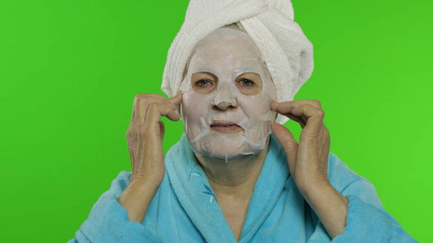 Avó idosa após o banho. Mulher velha aplicando máscara facial de tecido cosmético
 - Foto, Imagem