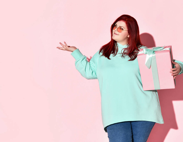 Young overweight woman in jeans, blue hoodie and sunglasses standing with gift box holding other hand up with open palm - 写真・画像