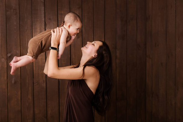 Young woman holding a newborn baby in her arms on the  brown background. Newborn photosession. Family portrait. Mother is kissing her baby. Baby goods packing template. Nursery. Medical and healthy. - Photo, Image