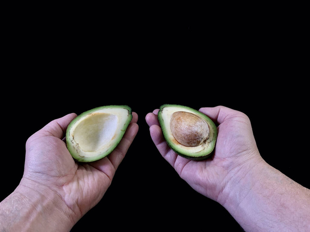 Halved avocado with a bone in his hand, on a black background. Green, ripe avocado vegetable in a cut. Halves Persea exotic fruit of an evergreen plant. - Photo, Image