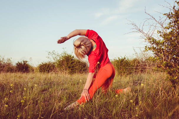 Young blond woman, wearing red leggings, doing evening exercises outside in summer. Fitness yoga stretching training outside. Healthy life concept. Active way of life - Photo, image