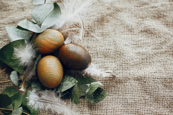 Κομψά πασχαλινά αυγά σε ρουστίκ φωλιά με φτερά στο ρουστίκ τραπέζι. Φυσικά βαμμένα πράσινα πασχαλινά αυγά με κλαδιά ευκαλύπτου, ανοιξιάτικα λουλούδια σε αγροτικό υφασμάτινο φόντο. Επίπεδη - Φωτογραφία, εικόνα