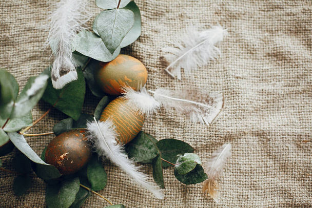 Stylish easter eggs in rustic nest with feathers on rustic table. Natural dyed green easter eggs with eucalyptus branch, spring flowers on rural textile background. Flat lay - Photo, Image