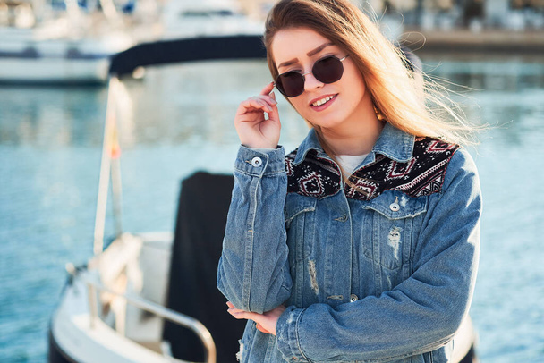 Beautiful girl holds round glasses wearing a denim jacket with long blonde hair on a summer day outdoors in the port - Fotoğraf, Görsel