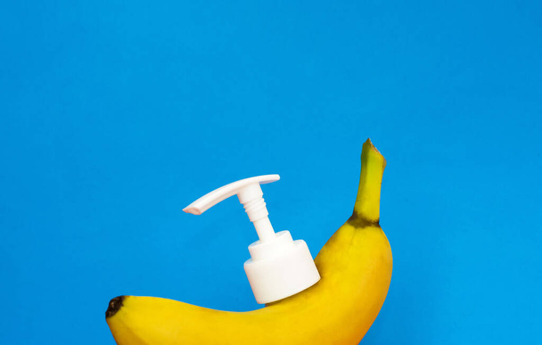 Banana with white dispenser for cosmetics, soap and other hygiene products on blue background. Useful exotic fruit. - Фото, изображение
