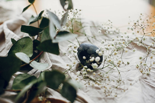 Happy Easter. Stylish easter egg in rustic nest with feathers on wooden table. Natural dyed blue easter egg with eucalyptus branch, white spring flowers on rural textile background. - Fotografie, Obrázek