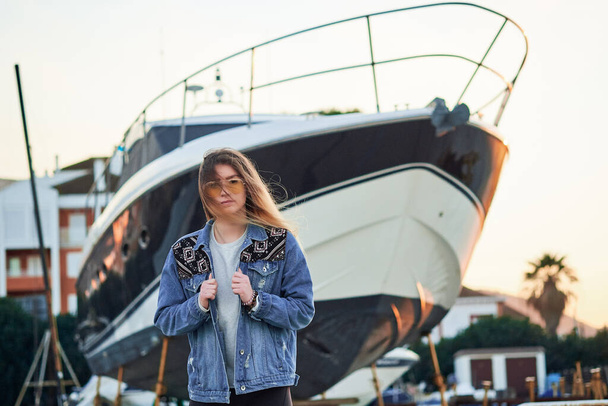 beautiful girl in sunglasses and a denim jacket with long blonde hair in a summer sunset on the background of a large luxurious yacht - 写真・画像