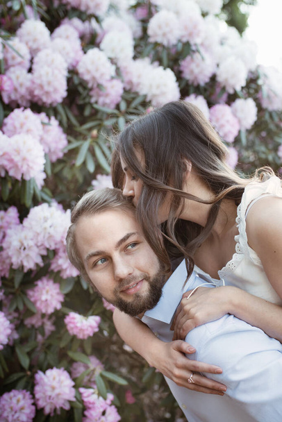 Emotioneel liefdesverhaal van een jong stel. Vrouw zit op de schouders van een man in een rhododendrons park in Engeland achter roze bloeiende struik, boom. De vrouw draagt een wit topje, de man draagt een lichtblauw shirt. Vrouw kust haar man - Foto, afbeelding