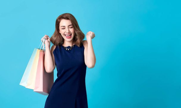 Portrait of an excited beautiful woman wearing blue dress and holding shopping bags isolated on blue background. - Photo, Image
