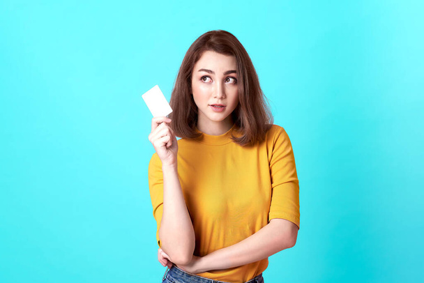 Jonge glimlachende mooie vrouw in geel shirt tonen credit card in de hand over blauwe achtergrond. - Foto, afbeelding