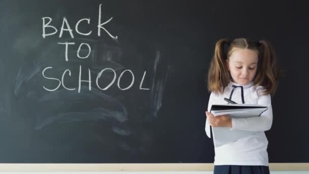 Caucasian girl student stands near the blackboard and writes text in a notebook. The inscription on the blackboard. Back to school concept - Footage, Video