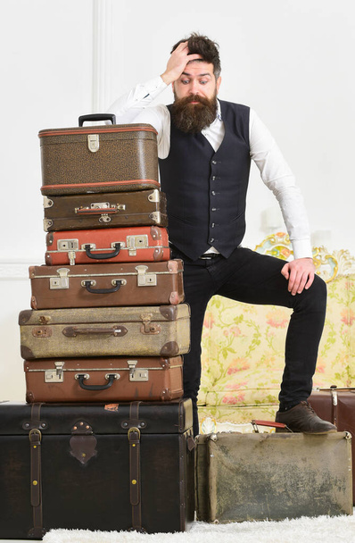 Macho elegant on disappointed face standing near pile of vintage suitcase. Luggage and travelling concept. Man, butler with beard and mustache delivers luggage, luxury white interior background - Photo, image