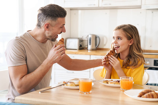 Photo of funny caucasian father and daughter laughing while having breakfast in modern kitchen - Foto, Imagem