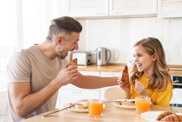 Photo of funny caucasian father and daughter laughing while having breakfast in modern kitchen - Foto, afbeelding