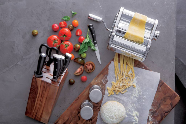 Ιταλική μακαρονάδα, μηχανή ζυμαρικών, μαγειρικά ζυμαρικά με ντομάτες και μαχαίρια, θέα από ψηλά. Σπιτική σπεσιαλιτέ Ιταλικά ζυμαρικά με υλικά για παραδοσιακή μεσογειακή κουζίνα σε τραπέζι κουζίνας - Φωτογραφία, εικόνα