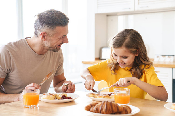 Photo of funny caucasian father and daughter laughing while having breakfast in modern kitchen - Фото, зображення