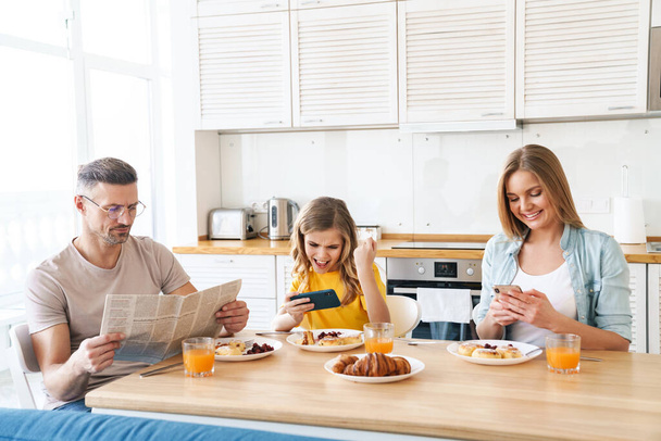 Kuva onnellisesta valkoihoisesta perheestä, joka käyttää älypuhelimia ja lukee sanomalehteä aamiaisen aikana modernissa keittiössä
 - Valokuva, kuva