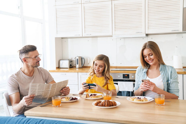 Foto da família caucasiana feliz usando smartphones e ler jornal enquanto toma café da manhã na cozinha moderna
 - Foto, Imagem