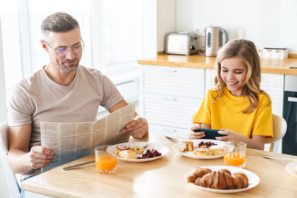 Photo of funny caucasian father and daughter using cellphone and reading newspaper while having breakfast - Photo, Image