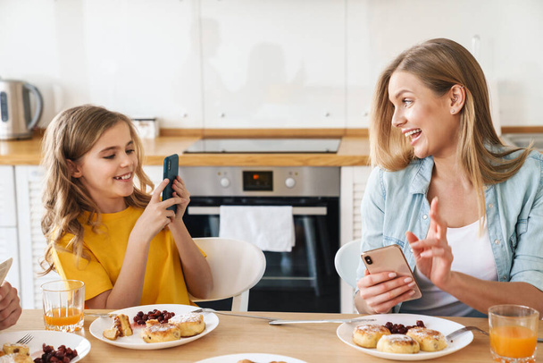 Photo of pleased caucasian mother and daughter taking selfie on cellphone while having breakfast in modern kitchen - Photo, Image
