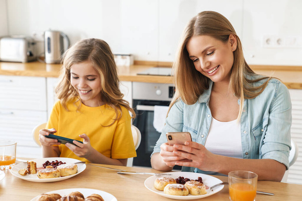 Photo of pleased caucasian mother and daughter using cellphones while having breakfast in modern kitchen - 写真・画像