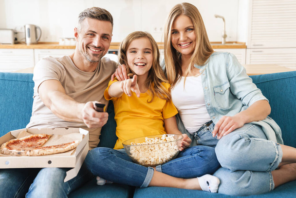 Photo of cheerful beautiful caucasian family eating popcorn and pizza while watching tv at home - Фото, изображение