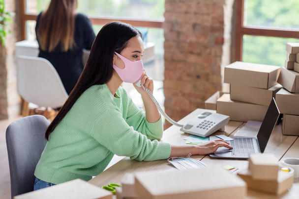 Profile photo of chinese business lady manager organize facial medical masks orders delivery boxes speak landline customer details noting information notebook home office indoors - Foto, imagen