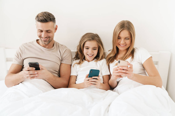 Photo of joyful caucasian family smiling and using cellphones while sitting in white bad after sleep - Φωτογραφία, εικόνα