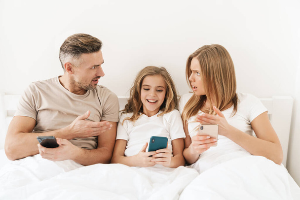 Photo of young caucasian family expressing shock and using cellphones while sitting in white bad after sleep - Foto, Imagem