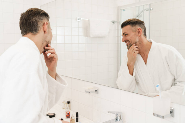 Photo of pleased caucasian man wearing white bathrobes smiling and looking at mirror in bathroom - Fotó, kép
