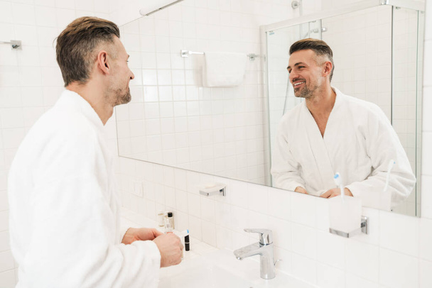 Beyaz bornoz giyen, elinde diş fırçası tutan ve banyoda aynaya bakan beyaz bir beyaz adamın fotoğrafı. - Fotoğraf, Görsel