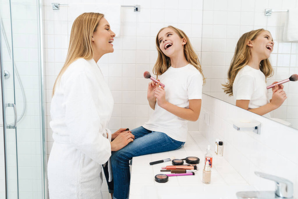 Photo of joyful caucasian mother and daughter smiling while doing makeup in white bathroom - Foto, afbeelding