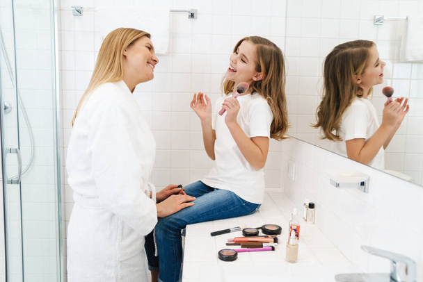 Photo of joyful caucasian mother and daughter smiling while doing makeup in white bathroom - Fotografie, Obrázek