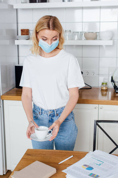 young woman in medical mask holding saucer and cup of coffee  - Photo, Image
