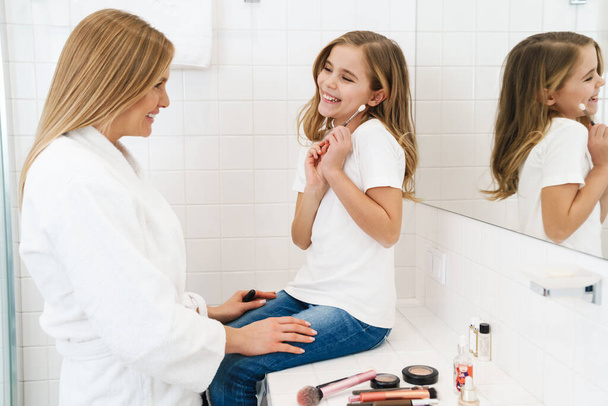 Photo of joyful caucasian mother and daughter smiling while doing makeup in white bathroom - Фото, изображение