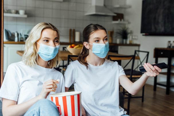 Sisters in medical masks holding popcorn while watching tv at home  - Fotografie, Obrázek